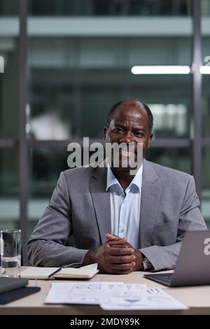 Erfolgreicher, reifer, männlicher Geschäftsführer eines afroamerikanischen Unternehmens, der auf die Kamera schaut, während er am Arbeitsplatz mit dem Computer sitzt Stockfoto