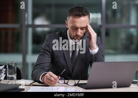 Gestresster männlicher Geschäftsführer, der den Kopf mit der Hand berührt und sich mit dem Laptop über den Arbeitsplatz bückt, während er Überstunden im Büro macht Stockfoto