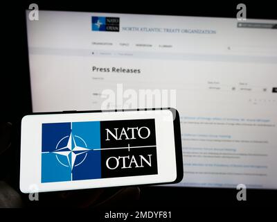 Person, die ein Mobiltelefon mit dem Logo der NATO (North Atlantic Treaty Organization) auf dem Bildschirm vor der Webseite hält. Konzentrieren Sie sich auf das Display des Telefons. Stockfoto