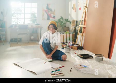 Junger Künstler in Freizeitkleidung sitzt auf dem Boden des Heimstudios und sieht sich leeres Papier an, während er sich für die Gestaltung neuer Kunstwerke inspirieren lässt Stockfoto