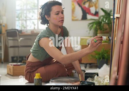 Junge, seriöse Frau in Freizeitkleidung, die während der Erstellung neuer Kunstwerke einen Pinsel hält, während sie im Studio auf dem Boden sitzt Stockfoto