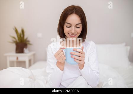Glückliche junge Frau, die zu Hause Kaffee im Bett trinkt Stockfoto