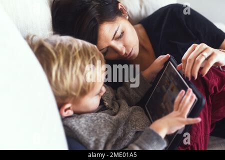 Mami schläft ein. Eine sorgenfreie junge Frau, die tagsüber mit ihrem kleinen Jungen auf einem digitalen Tablet surft, während sie zu Hause auf dem Sofa sitzt. Stockfoto