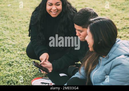 Eine Gruppe von Latinos, die lachten, saßen auf dem Boden in einem Park mit einem Smartphone Stockfoto