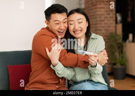 Familien- und Beziehungskonzept. Glückliches schwangeres asiatisches Paar mit Schwangerschaftstest, das umarmt und lächelt, auf der Couch sitzt Stockfoto