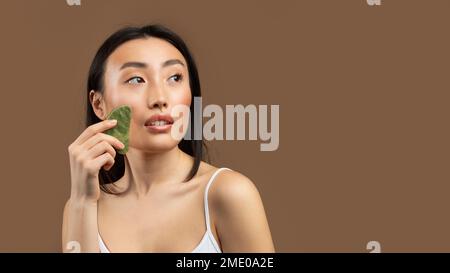 Junge japanische Dame, die Jade-Massage mit Gua-Sha-Stein macht, auf braunem Hintergrund posiert, Panorama mit freiem Platz Stockfoto