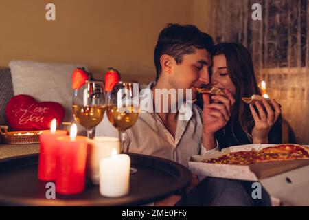 Valentinstagspaar, das Pizza isst, Champagner trinkt, ein romantisches Abendessen zu Hause feiert Stockfoto