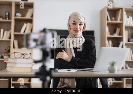 Eine wunderschöne muslimische Frau im Hijab, die spricht, während sie Videos auf einem modernen Smartphone ansieht und aufnimmt. Weibliche Bloggerin, die zu Hause sitzt und Livestream macht. Stockfoto