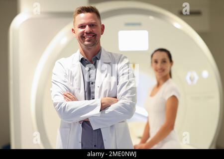 Selbstbewusste, lächelnde männliche Ärztin aus der Nahaufnahme und Patientin und CT-Scanner im Hintergrund. Stockfoto