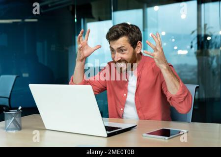 Wütender und nervöser Geschäftsmann, der per Fernzugriff per Videoanruf telefoniert, Mann, der Kollegen online anschreit, Chef im Hemd, der in einem modernen Büro mit einem Laptop arbeitet. Stockfoto