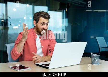 Wütender und nervöser Geschäftsmann, der per Fernzugriff per Videoanruf telefoniert, Mann, der Kollegen online anschreit, Chef im Hemd, der in einem modernen Büro mit einem Laptop arbeitet. Stockfoto