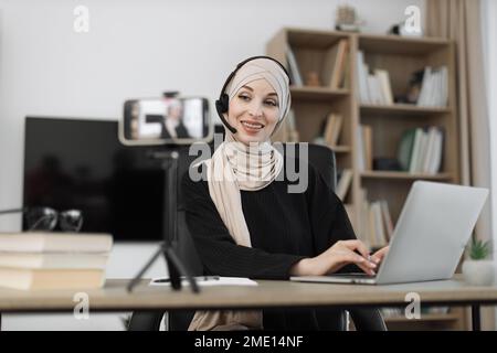 Selbstbewusste muslimische Frau mit Kopftuch und Headset sitzt am Schreibtisch und arbeitet an einem Laptop und nimmt Videos auf einem digitalen Smartphone auf. Freiberufler-Konzept und moderne Technik. Stockfoto