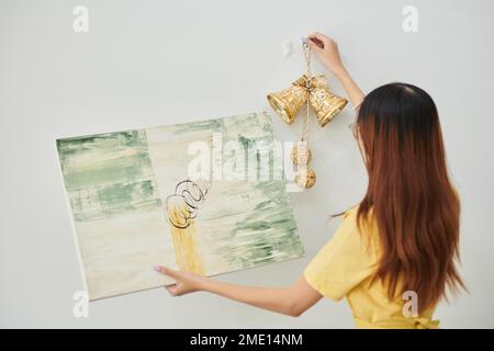 Junge Frau, die goldene Glocken aufhängt und ein abstraktes Bild an der Wand in der Wohnung, Blick von hinten Stockfoto