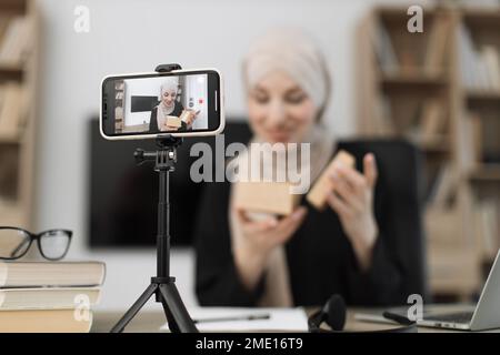 Konzentrieren Sie sich auf das Telefon einer glücklichen muslimischen Bloggerin, die Geschenkboxen von Unternehmen auspackt und Live-Streaming durchführt. Arabische Frau, die Smartphone-Kamera benutzt, um Inhalte zu erstellen. Stockfoto