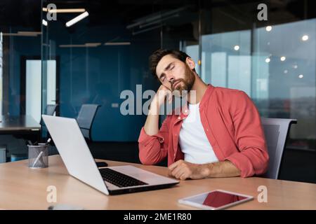 Müder Geschäftsmann im lässigen roten Hemd schläft am Arbeitsplatz, der Mann am Schreibtisch schlief während der Arbeitszeit im Büro mit einem Laptop ein. Stockfoto