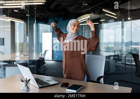 Junge arabische Geschäftsfrau in Hijab tanzt im Büro neben dem Tisch, hört Musik auf einem Laptop. Entspannt und lächelnd hob sie ihre Hände. Ruhe, Pause. Stockfoto