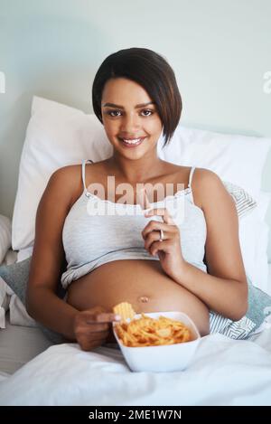 Das Baby will, was es will. Porträt einer attraktiven jungen schwangeren Frau, die Kartoffelchips und Schokolade im Bett isst. Stockfoto