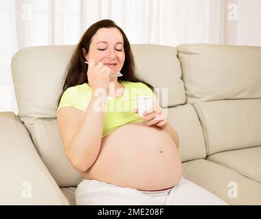 Eine schwangere Frau, die im Wohnzimmer sitzt und Joghurt isst Stockfoto