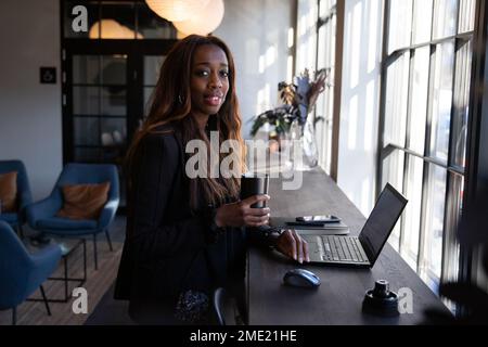 Lächelnde afroamerikanische Geschäftsfrau, die im Büro Kaffee trinkt und mit einem Laptop arbeitet Stockfoto