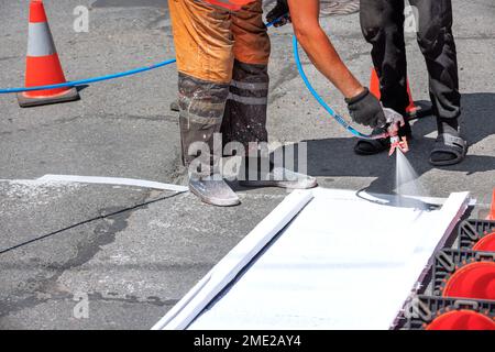 Ein Straßenarbeiter in schmutzigen Overalls bringt an einem Sommertag mit einer Spritzpistole weiße Straßenmarkierungen auf dem Asphaltbelag an. Speicherplatz kopieren. Stockfoto