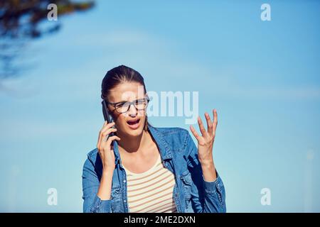Woher hast du diese Nummer? Eine attraktive junge Frau, die draußen ein Handy benutzt und wütend aussieht. Stockfoto