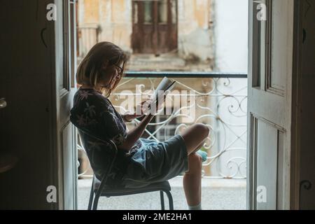 Seitenansicht einer Frau in zwangloser Kleidung, die auf einem Stuhl neben offenen Balkontüren sitzt und interessante Romane in Kuba liest Stockfoto