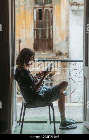 Seitenansicht einer Frau in zwangloser Kleidung, die auf einem Stuhl neben offenen Balkontüren sitzt und interessante Romane in Kuba liest Stockfoto