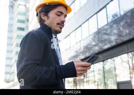 Von unten: Junger selbstbewusster männlicher Ingenieur in eleganter Freizeitkleidung mit dunklem Haar und gelbem Schutzhelm, der im Stehen ein Smartphone verwendet Stockfoto