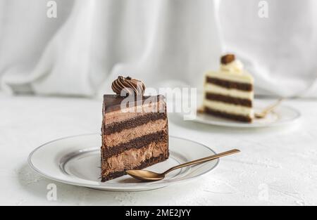 Ein Stück leckerer süßer Schokoladenkuchen und weißer Kuchen mit Glasur und Schlagsahne, serviert auf Keramikteller mit goldenem Löffel auf weißer Tischdecke Stockfoto