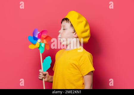 Ein Junge mit gelbem T-Shirt und Hut steht vor pinkfarbenem Hintergrund und bläst ein bunter Windrad Stockfoto