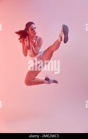 Der ganze Körper einer entschlossenen jungen Kämpferin mit Pferdeschwanz in farbenfroher Sportbekleidung führt im Studio einen Springkick auf pinkfarbenem Hintergrund auf Stockfoto