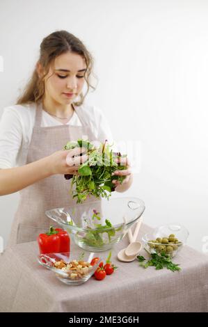 Das Mädchen mischt Salat mit den Händen Sie gießt ihn in eine Glasplatte vegetarisches Essen gesundes Essen Holzlöffel Schürze Tischdecke junge Frau Teenager bereitet Abendessen für die Familie zu Frühstück Mittagessen Stockfoto