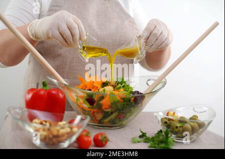 Kochen in sterilen Handschuhen bereitet köstliche gesunde Speisen zu, gießt Dressing in Salat in Glasschüssel aus zwei Kannen Salat Olivenöl und Soße mit Senf und Honigzutaten Stockfoto