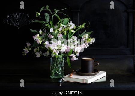 Sweetpea-Blumen im Glasgefäß bei Kaffeetasse und Tagebuch auf schwarzem Klavier Stockfoto