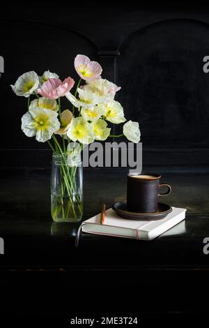 Frische isländische Mohnblumen in einem Glasgefäß mit Kaffeetasse und Tagebuch auf schwarzem Klavier Stockfoto