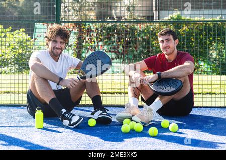 Freunde sitzen mit Tennisschlägern und Bällen auf dem Sportplatz Stockfoto