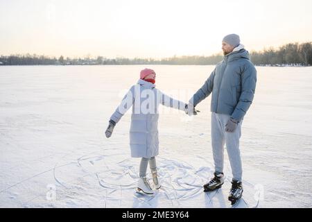 Vater hält die Hand der Tochter und übt Eislaufen auf dem Wintersee Stockfoto