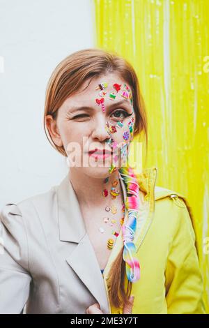 Frau mit Aufklebern im Gesicht, die vor einer zweifarbigen Wand zwinkert Stockfoto