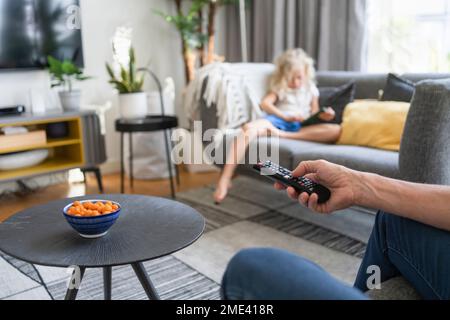 Hand eines Mannes mit Fernbedienung und Tochter im Hintergrund auf dem Sofa Stockfoto