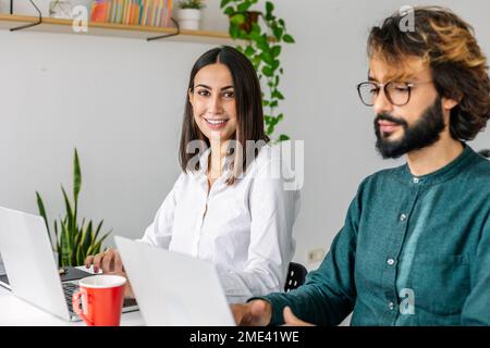 Lächelnde junge Geschäftsfrau von einem Kollegen, der am Schreibtisch ein Notebook benutzt Stockfoto