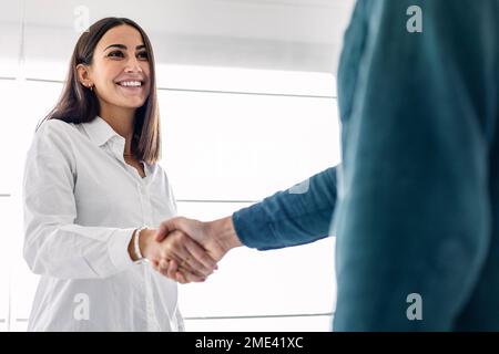 Glückliche junge Geschäftsfrau, die mit einem Kollegen am Arbeitsplatz die Hand schüttelt Stockfoto