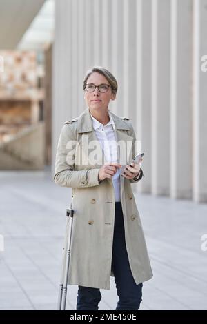 Reife Geschäftsfrau mit Rollgepäck, die in der Stadt steht und ein Smartphone hält Stockfoto