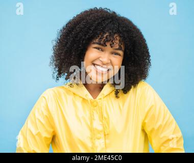 Frau, lächeln und glücklich im Porträt mit Mode für den Winter, Regenmantel und Schönheit isoliert auf blauem Hintergrund. Afrikaner, Glück im Studio und Stockfoto