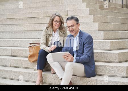 Ein erfahrener Geschäftsmann, der einen Tablet-PC verwendet, sitzt von einem Kollegen auf einer Treppe Stockfoto