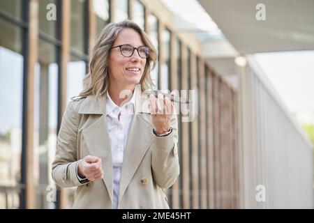 Lächelnde, reife Geschäftsfrau, die sich vor dem Gebäude mit einem Freisprechtelefon unterhält Stockfoto