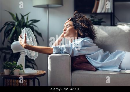 Junge Frau, die sich mit einem elektrischen Ventilator abkühlt und zu Hause auf dem Sofa liegt Stockfoto