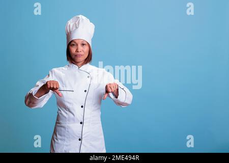 Ältere Köchin in Küchen-Uniform, die Missbilligung unterschreibt, nachdem sie sich ein kulinarisches Abendessen angesehen hat. Ein fröhlicher Koch, der mit dem Daumen nach unten zeigt und ein gesundes Gericht zubereitet Stockfoto
