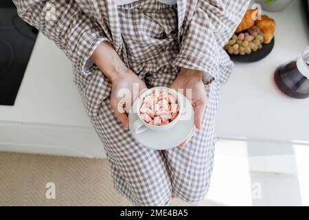 Hände einer Frau, die heiße Schokolade mit Marshmallows in der Tasse hält und auf der Küchentheke sitzt Stockfoto