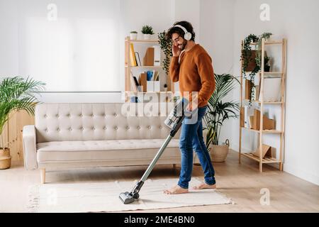 Glücklicher junger Mann mit kabellosen Kopfhörern, der sein Zuhause mit einem Staubsauger reinigt Stockfoto