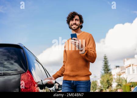 Glücklicher junger Mann mit Handy-Ladewagen an der Ladestation für Elektrofahrzeuge Stockfoto